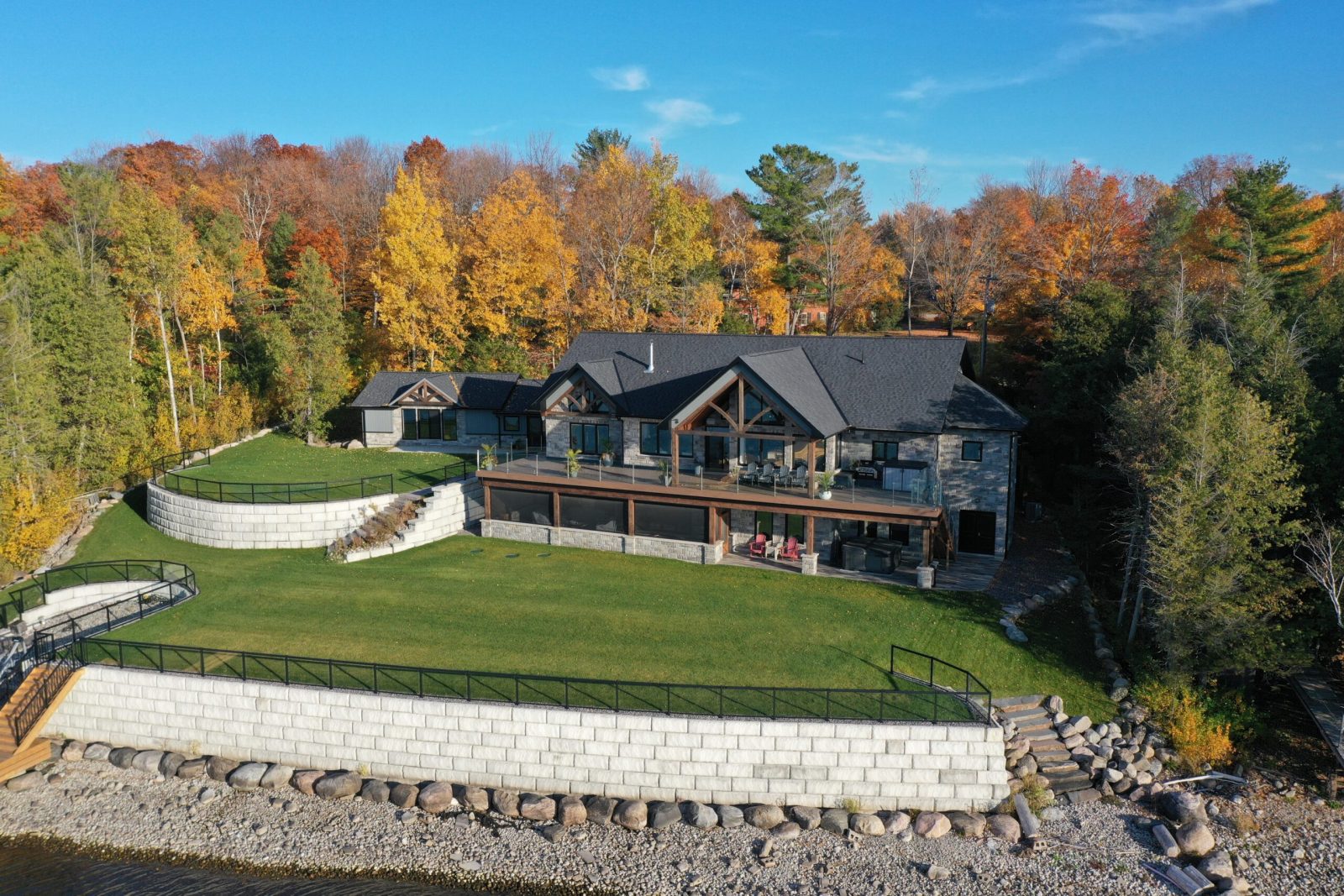 Elimere Point Custom Home - Aerial view of a large lakeside house surrounded by autumn trees. The house features dark roofing and stone walls, with expansive windows and multiple decks. A well-kept green lawn and a stone retaining wall lead down to the water’s edge.