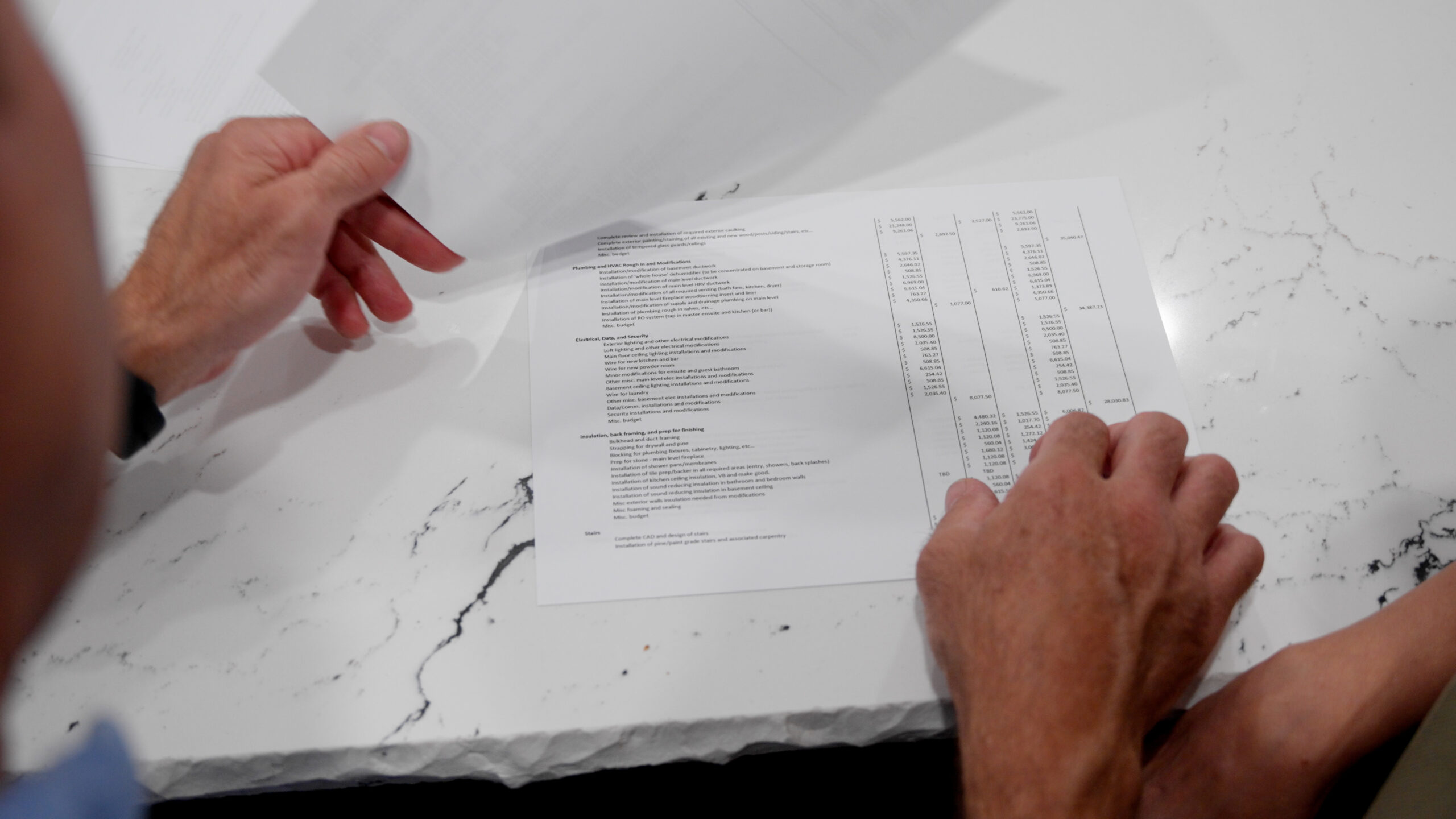 The Building Contract - Hands of a person holding and examining a document with text and charts on a white marble table. The document has various sections and a vertical layout with numbered lines or lists. Another piece of paper is partially visible being lifted.