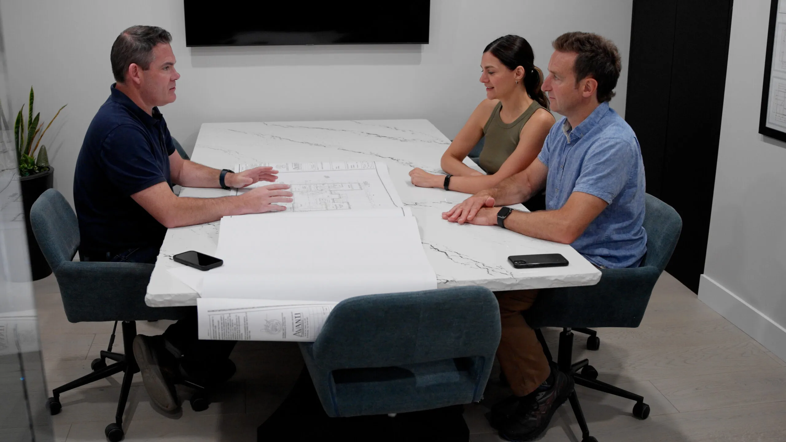 Three people sit around a white marble conference table, discussing a set of architectural drawings. A man gestures towards the plans while the others attentively listen. A plant, smartphone, and wall-mounted screen are visible in the room.