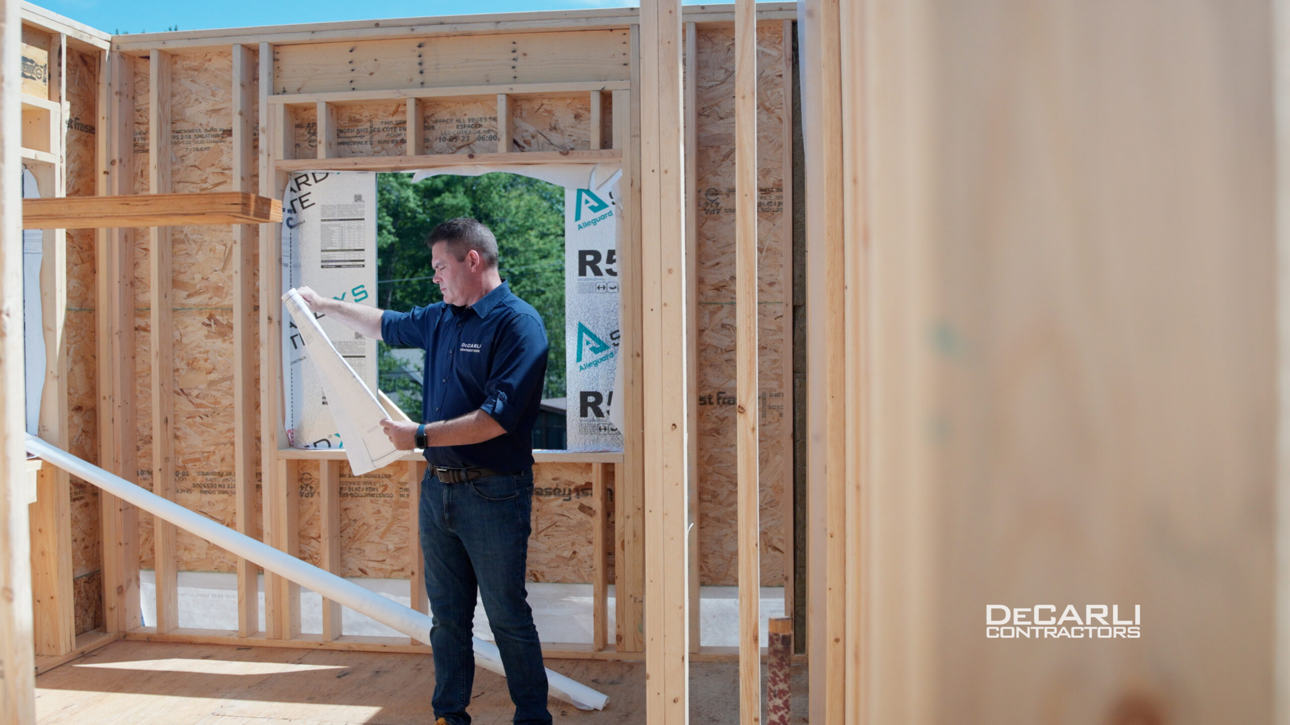 A person in a blue shirt and jeans examines a blueprint inside the wooden frame of a house under construction. Sunlight illuminates the site, showcasing wooden beams and an opening with a partially covered window. DeCarli Contractors is visible in the corner.