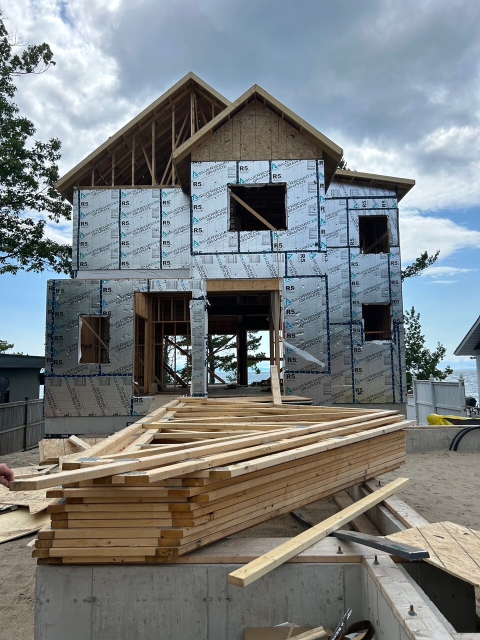 A partially constructed two-story house with visible plywood and metallic insulation panels. Wooden planks are stacked in the foreground. The sky is cloudy, and trees are visible on the left.