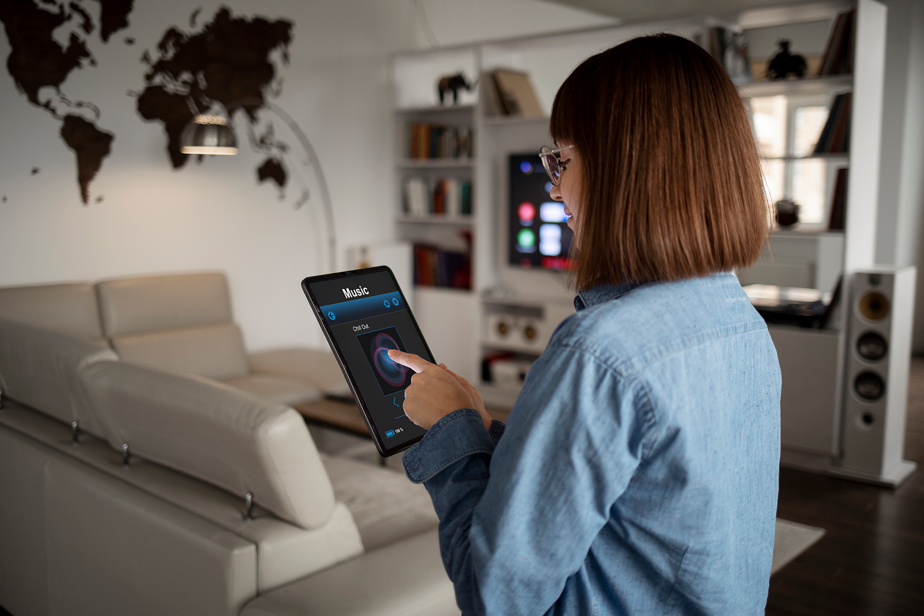 A person wearing glasses stands in a modern living room, using a tablet to control a music app. The room features a world map on the wall, a beige couch, and a TV with speakers.