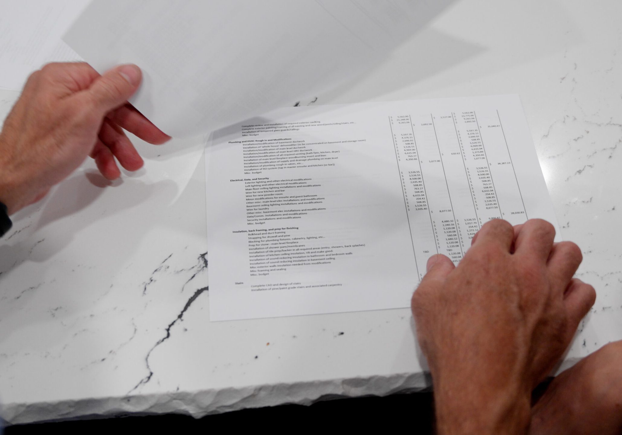 The Building Contract - Hands of a person holding and examining a document with text and charts on a white marble table. The document has various sections and a vertical layout with numbered lines or lists. Another piece of paper is partially visible being lifted.