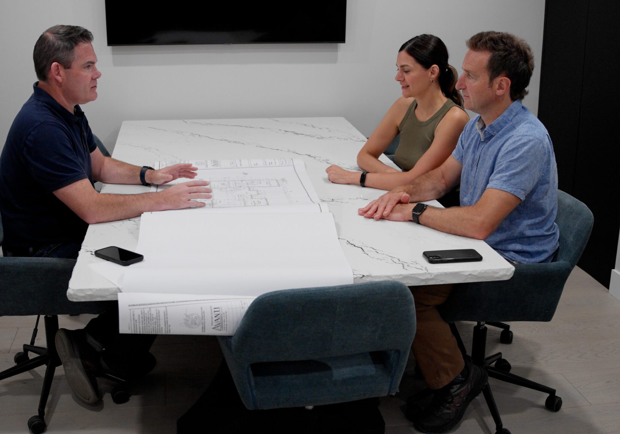 Three people sit around a white marble conference table, discussing a set of architectural drawings. A man gestures towards the plans while the others attentively listen. A plant, smartphone, and wall-mounted screen are visible in the room.