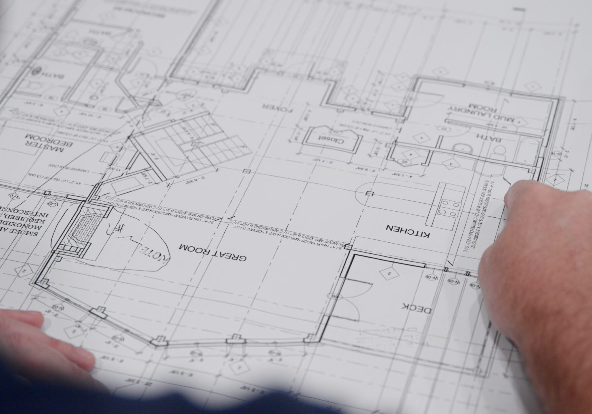 Close-up of a person reviewing architectural blueprints. The plans detail a building layout including spaces labeled Kitchen, Great Room, and Deck. The person is pointing at the blueprint, which is detailed with measurements and notes.