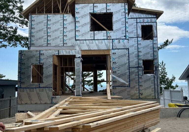 A partially constructed two-story house with visible plywood and metallic insulation panels. Wooden planks are stacked in the foreground. The sky is cloudy, and trees are visible on the left.