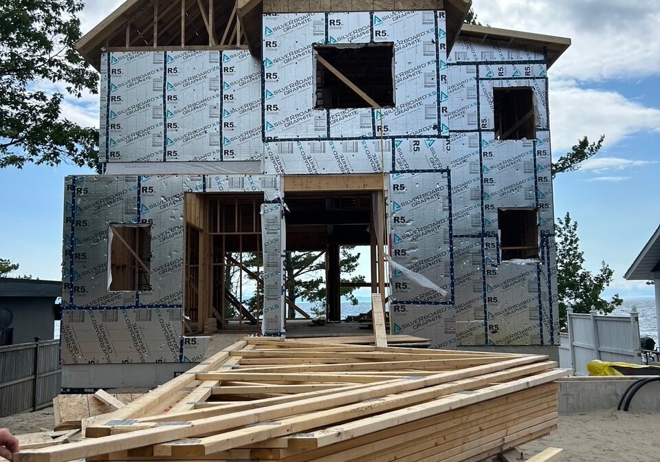A partially constructed two-story house with visible plywood and metallic insulation panels. Wooden planks are stacked in the foreground. The sky is cloudy, and trees are visible on the left.