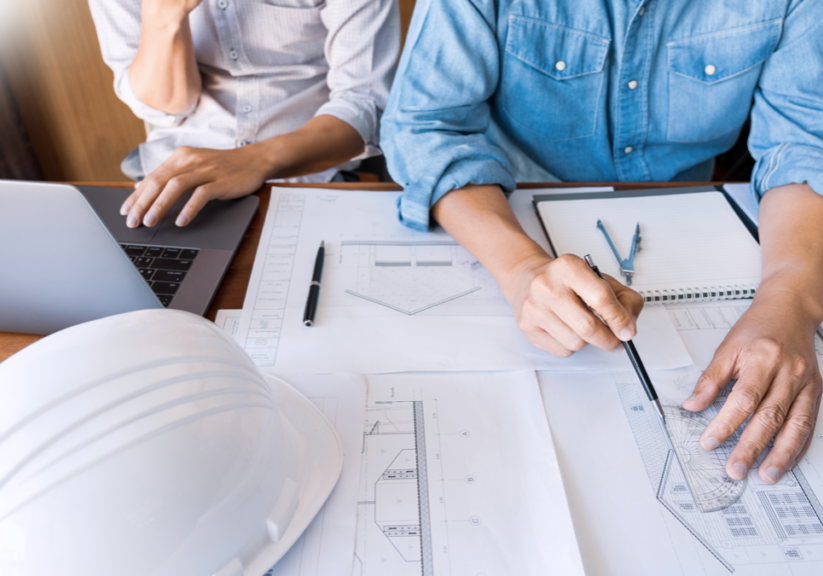 Detailed Quotations - Two people work at a desk with architectural plans, a laptop, notepad, and a white safety helmet. One person points at the plans with a pen while the other looks at the laptop screen. Both are wearing long-sleeved shirts.