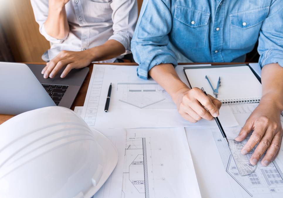Two people work at a desk with architectural plans, a laptop, notepad, and a white safety helmet. One person points at the plans with a pen while the other looks at the laptop screen. Both are wearing long-sleeved shirts. Detailed Quotations - Two people work at a desk with architectural plans, a laptop, notepad, and a white safety helmet. One person points at the plans with a pen while the other looks at the laptop screen. Both are wearing long-sleeved shirts.