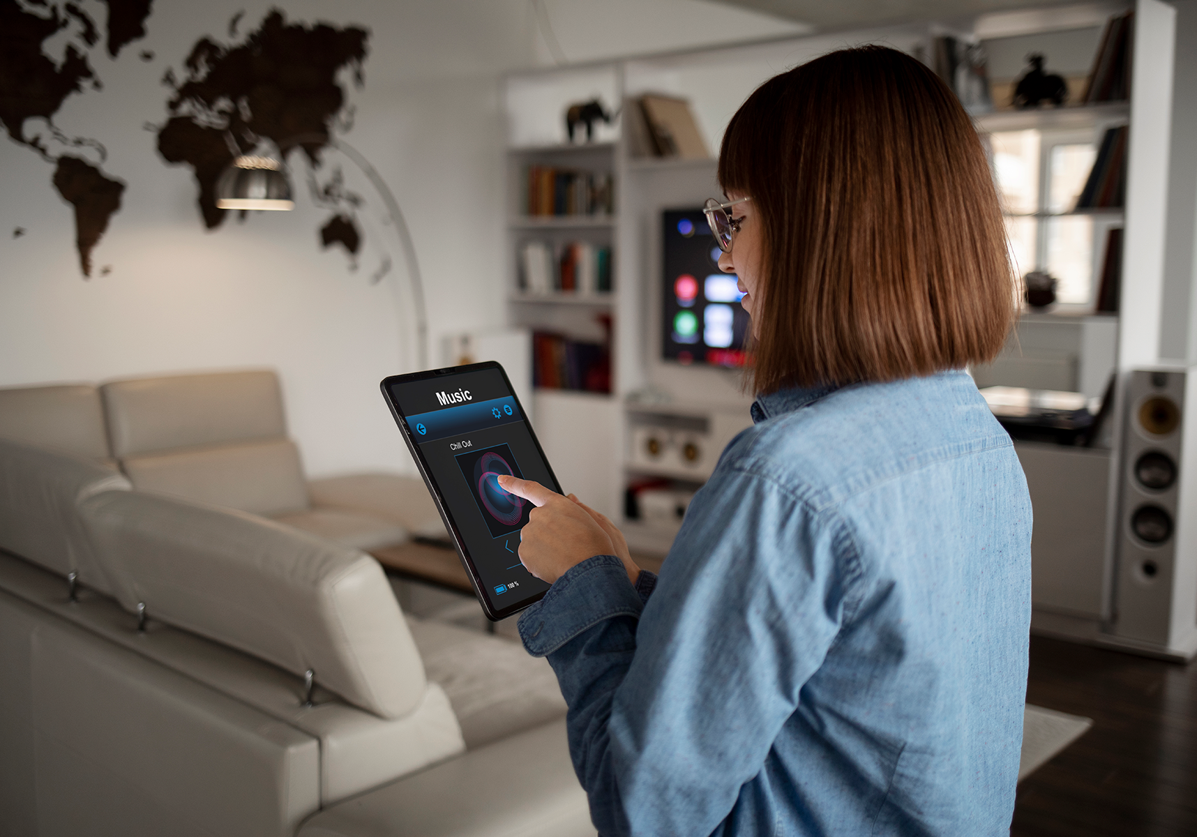 A person wearing glasses stands in a modern living room, using a tablet to control a music app. The room features a world map on the wall, a beige couch, and a TV with speakers.